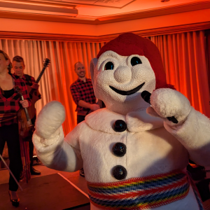 A person in a friendly snowman costume with a red scarf holds a microphone, performing on stage with musicians in the background. The scene is warmly lit with red and orange tones.