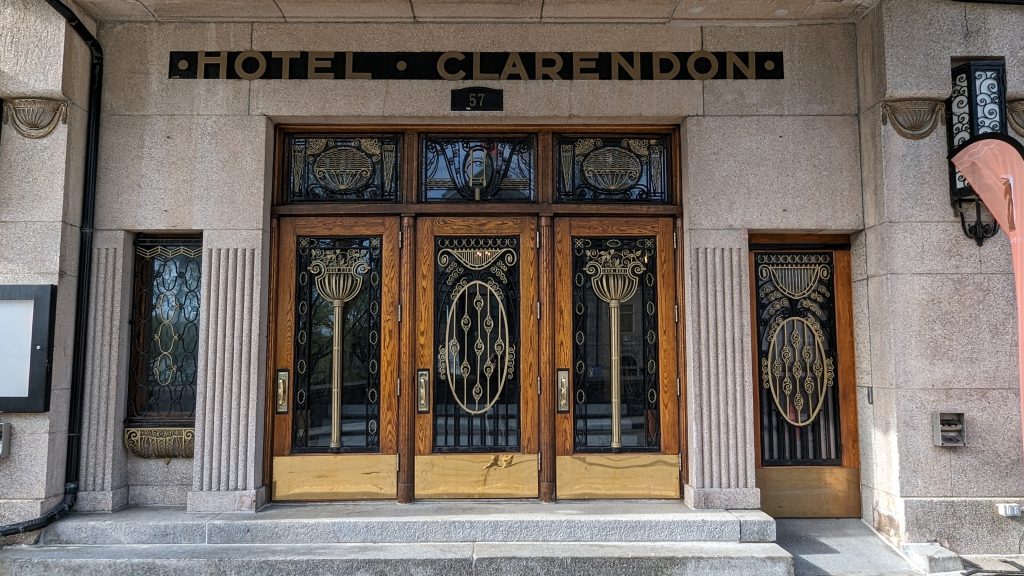 The entrance of Hotel Clarendon, near Hotel Le Vincent, features three wooden doors with intricate art deco glass designs, flanked by two matching side panels, and the hotel name displayed in bold letters above.
