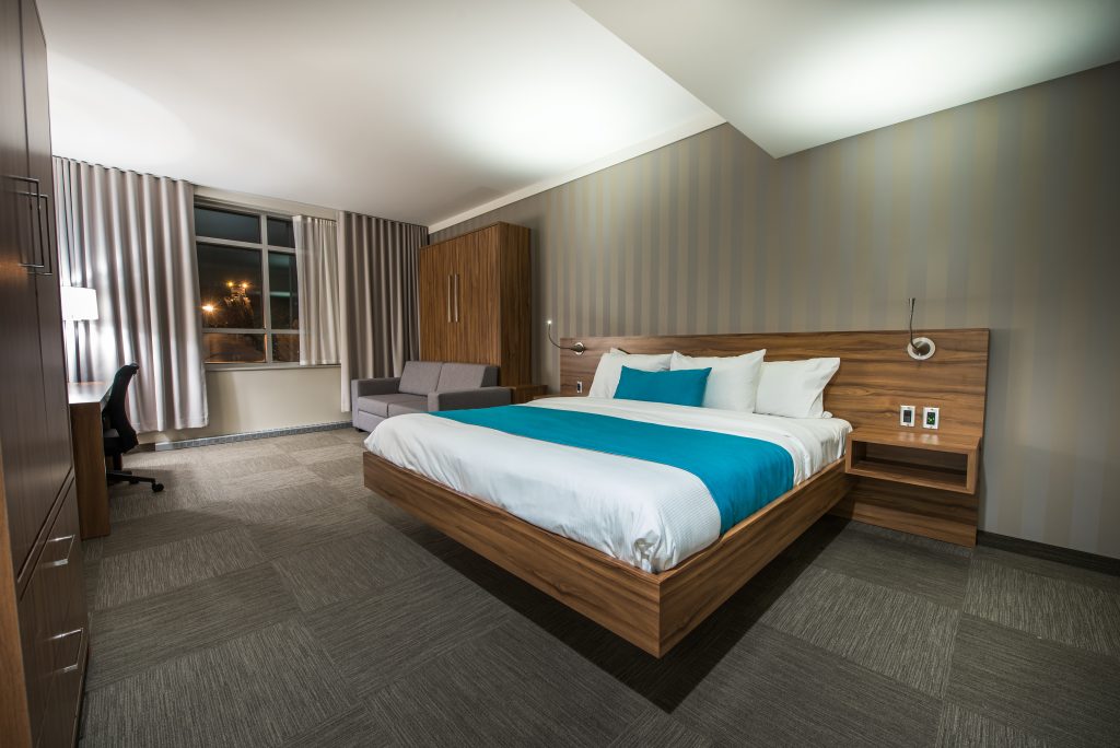Modern Hotel Clarendon room with a wooden bed frame, white bedding, and a teal runner. The space offers a sofa, desk, wardrobe, and large window with curtains. Neutral colors and soft lighting create a clean, inviting atmosphere.
