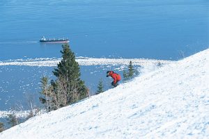 A person in a red jacket skis down a snowy slope near pine trees, with a cargo ship on a blue, partially frozen lake in the background—capturing the stunning winter scenery found at some of Canada’s best ski resorts.