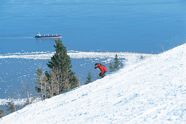 A person in a red jacket skis down a snowy slope near pine trees, with a cargo ship on a blue, partially frozen lake in the background—capturing the stunning winter scenery found at some of Canada’s best ski resorts.