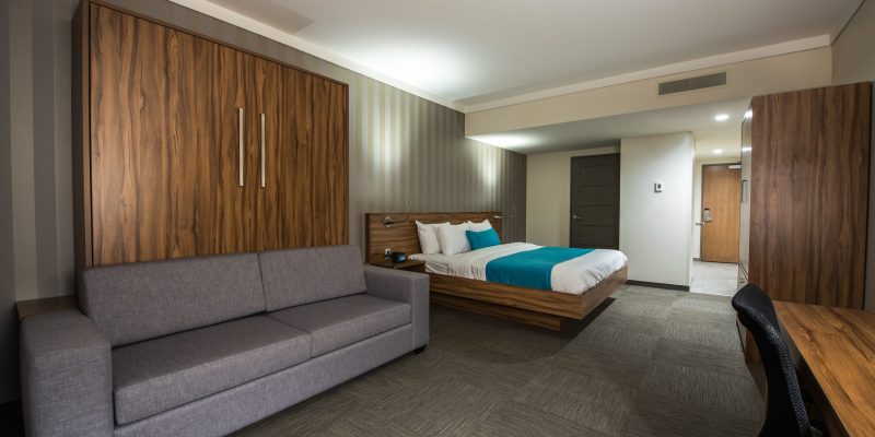 Modern Hotel Clarendon room with a gray sofa, wooden furniture, a bed with white and turquoise bedding, a desk and chair, plus neutral-colored walls and carpet. The space is well-lit and spacious for your comfort.