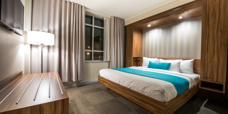 Modern hotel room at Hotel Clarendon with a double bed, blue runner and pillows, wood furnishings, a flat-screen TV, desk, and large window with curtains. The warm lighting creates a clean and inviting atmosphere.