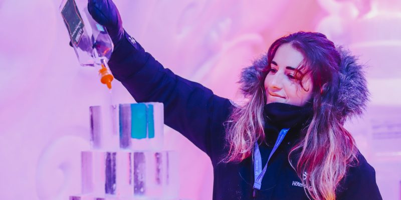 A woman in winter clothing and earmuffs pours liquid from a bottle onto a stack of clear ice blocks against a colorful, frosty background at Hotel Clarendon.