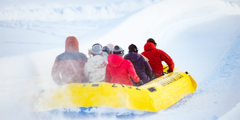 A group of people in winter clothing ride a yellow inflatable raft down a snowy slope near Hotel Clarendon, surrounded by snow-covered trees.